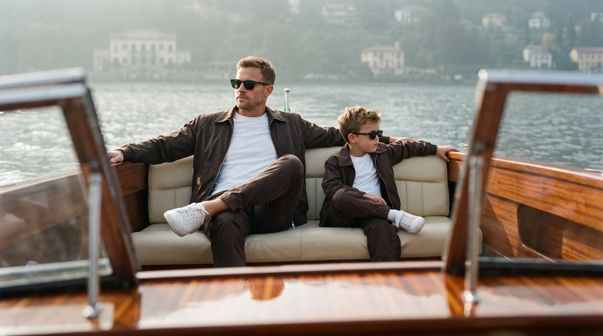 Father and son leaning back on a classic wooden boat on Lake Como in matching brown tech tracksuits