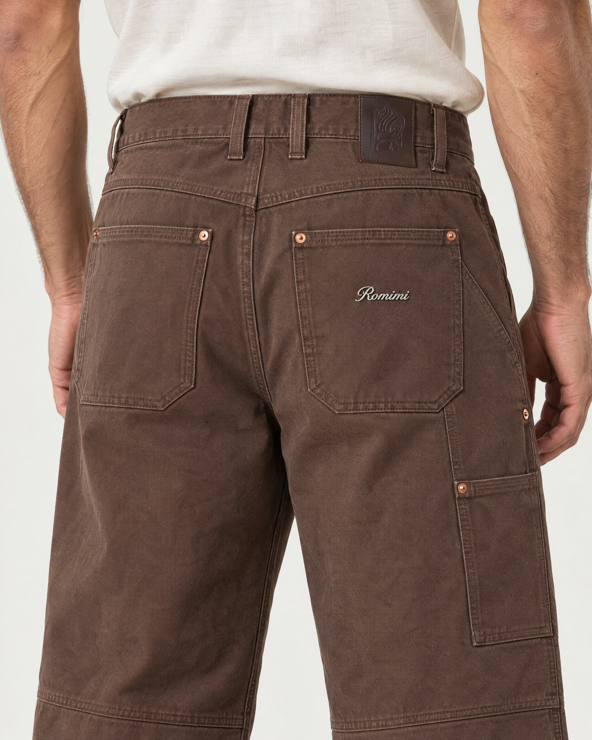 Detail of the Romimi carpenter pants in chocolate brown: the leather waist patch and silver metal Romimi logo on the back pocket